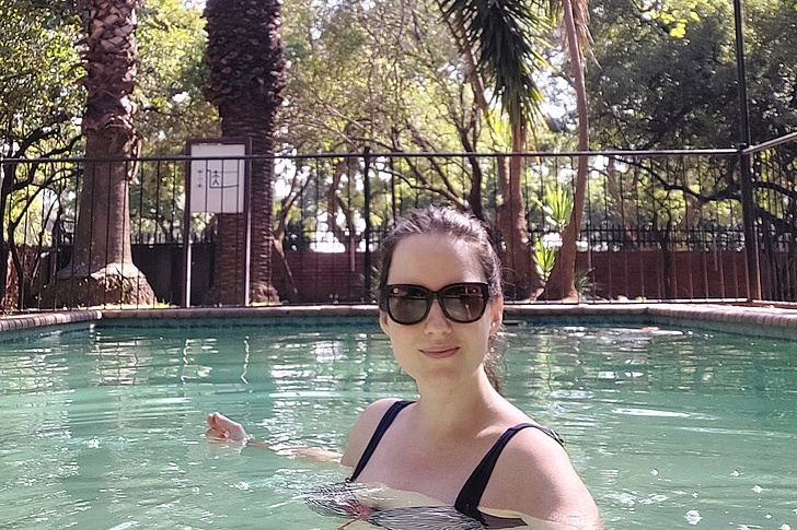 Person mit schwarzem Badeanzug entspannt im Schwimmbecken mit Umzäunung und Bäumen im Hintergrund