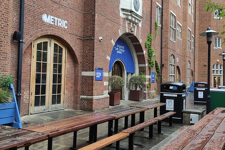 Eingang des Metric Imperial College in London mit Backsteinfassade, blauem Schild, Bänken und Pflanzen unter nassem Himmel
