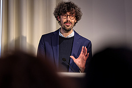 Zu sehen ist Dr. Romain Faure. Er steht vor einem beigefarbenen Vorhang und spricht an einem Rednerpult in ein Mikrofon. Er hebt seine linke Hand beim Sprechen.