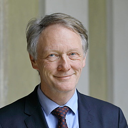 Middle-aged man with gray hair wearing a dark suit, light blue shirt, and patterned tie against a light background