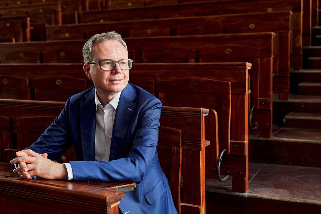 Andreas Reckwitz sitzt in einem alten Hörsaal auf einem Stuhl und hat die Hände und Arm auf der Ablage vor ihm gelegt. Er blickt an der Kamera vorbei nach rechts. Er trägt ein helles Hemd und einen royalblauen Anzug.