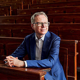 Andreas Reckwitz sitzt in einem alten Hörsaal auf einem Stuhl und hat die Hände und Arm auf der Ablage vor ihm gelegt. Er blickt an der Kamera vorbei nach rechts. Er trägt ein helles Hemd und einen royalblauen Anzug.