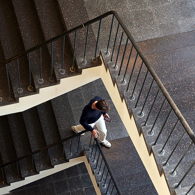 Campus Mitte Eine Person, die eine Wendeltreppe hinuntergeht. Die Treppe hat dunkle Stufen und ein Geländer mit vertikalen Stäben. Der Boden ist ebenfalls dunkel gefliest. Die Person trägt einen blauen Pullover, weiße Hosen und hält ein Handy in der Hand.