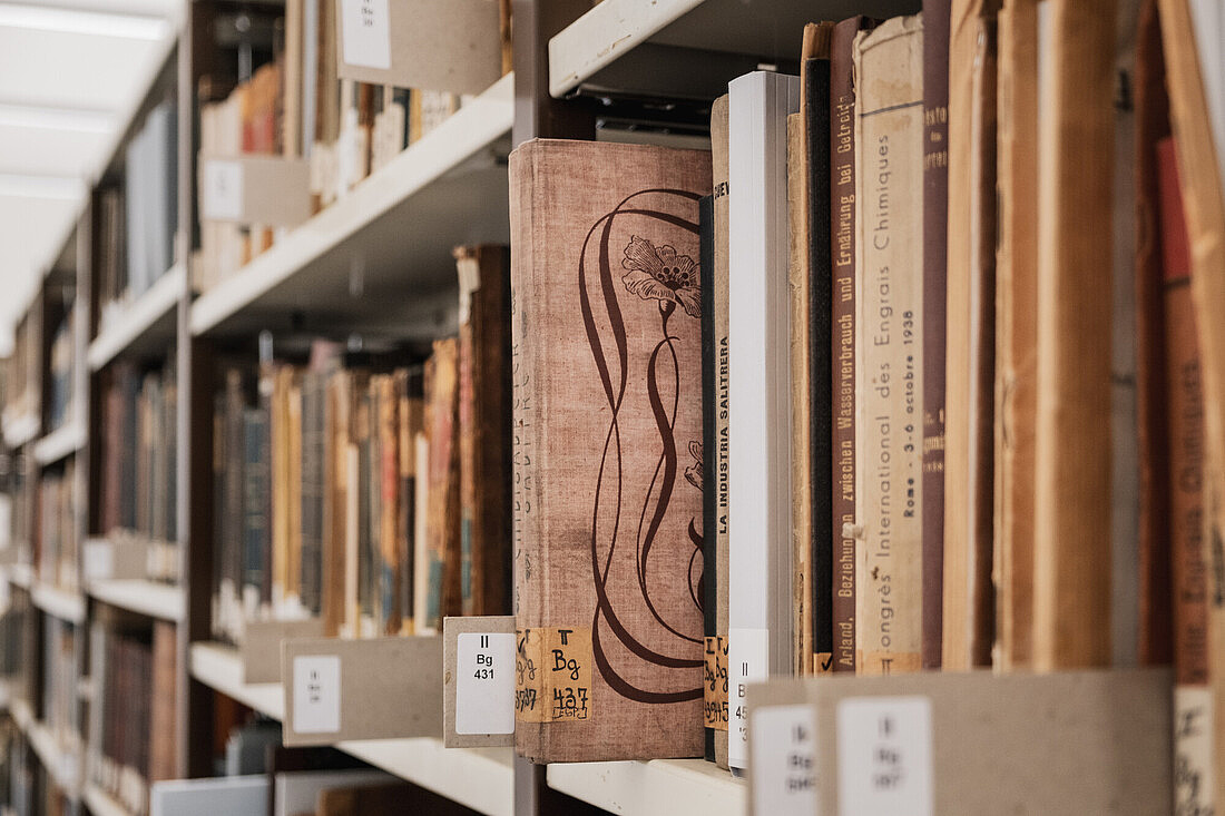 Campus Nord Eine Buchreihe mit sehr alt aussehenden Büchern in einer Bibliothek. Eines der Bücher ragt hervor. Darauf ist eine Zeichnung einer Blume zu sehen.