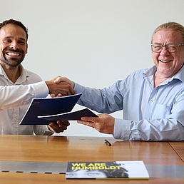 Two smiling men sit opposite each other, shaking hands and holding a file in their other hand.