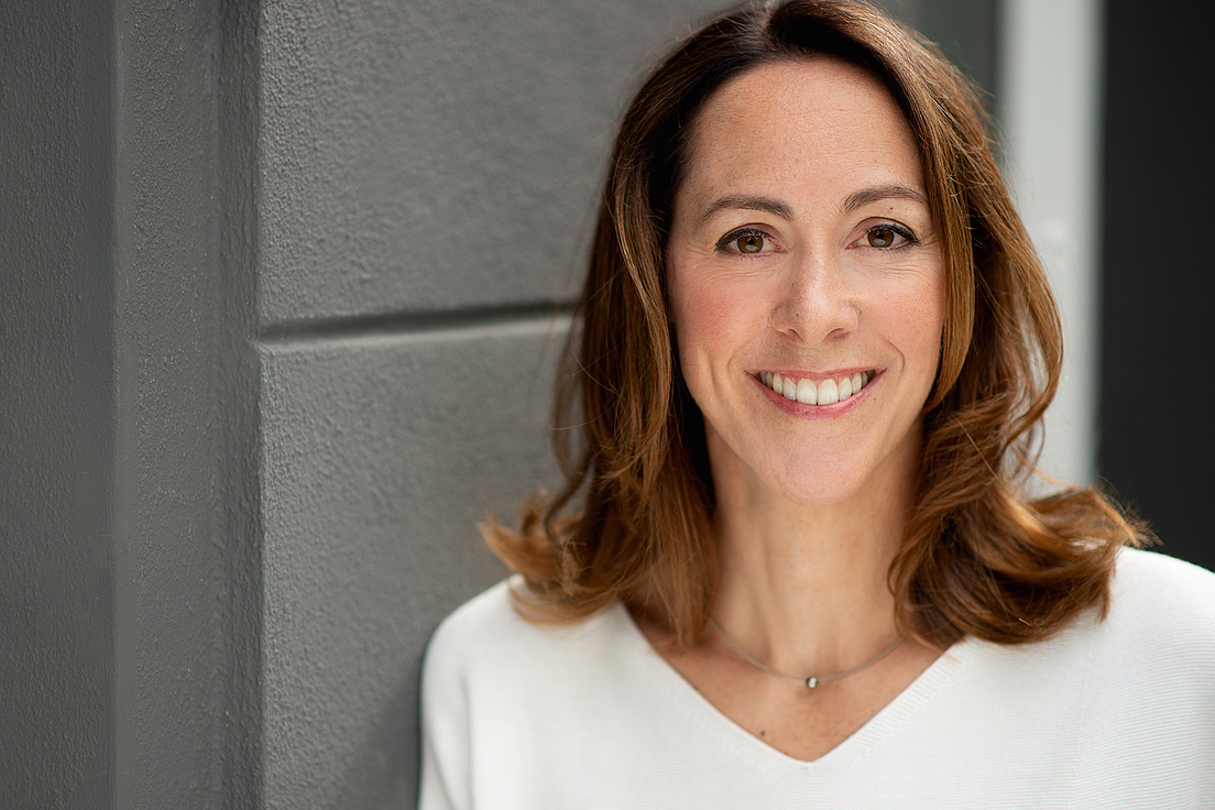 Gisela Rühl lächelt in die Kamera und trägt eine weiße Bluse und eine Kette mit einem Edelstein