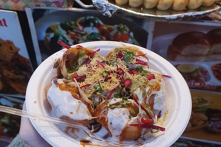 Schüssel Pani Puri: Fünf gefüllte Teigbällchen mit Joghurt, Chutney, Kichererbsen, Granatapfelkernen, Koriander und Radieschen, serviert auf weißem Pappteller mit Löffel.