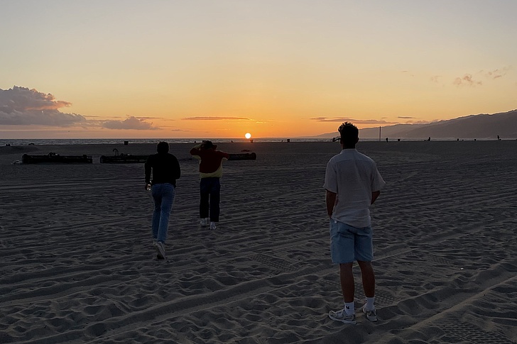 Drei Personen gehen auf einem Strand bei Sonnenuntergang, zwei von ihnen sind von hinten zu sehen, eine steht näher und trägt Shorts und ein T-Shirt