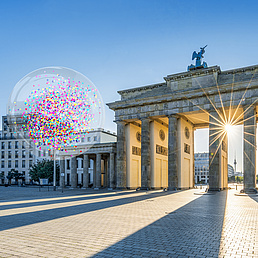 Ein Bild vom Brandenburger Tor, im Hintergrund blauer Himmel und Sonnenstrahlen, die zwischen den Säulen hervorscheinen. Im Bild sind auch vier transparente Blasen mit Konfetti zu sehen. 