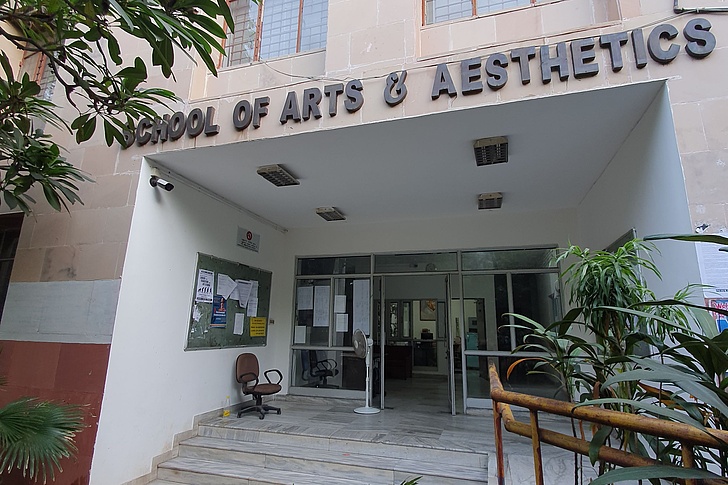 Eingang zur School of Arts & Aesthetics: Offene Glastüren mit Anschlagtafeln, Stuhl, Pflanzen und beleuchtete Veranda vor einem cremefarbenen Gebäude mit Palmen links.