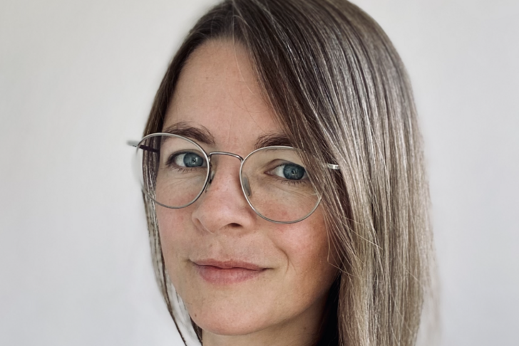 Nina Hanning mit braunen, langen Haaren und blauen Augen blickt direkt in die Kamera. Sie trägt eine runde Brille mit einem schmalen Metallgestell und ein weißes Hemd mit schwarzen Punkten. Ihr Haar fällt über ihre Schulter, und sie hat einen leichten, freundlichen Ausdruck im Gesicht. Der Hintergrund ist einheitlich weiß.