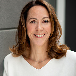 Gisela Rühl lächelt in die Kamera und trägt eine weiße Bluse und eine Kette mit einem Edelstein