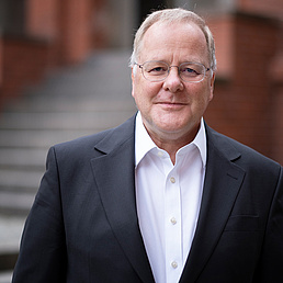 Georg Essen trägt ein weißes Hemd und einen dunkelfarbenen Anzug. Er steht vor dem Eingangsportal des Zentralinstituts für Katholische Theologie der HU. Es ist ein rotfarbenes Backsteingebäude.