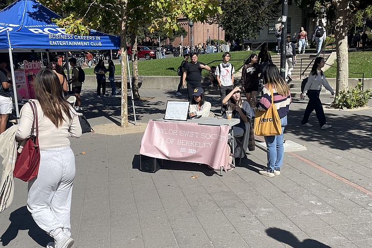 Menschen stehen und gehen auf einem sonnigen Campusweg, ein Stand mit einem rosa Banner 'Taylor Swift Society of Berkeley' ist aufgebaut.