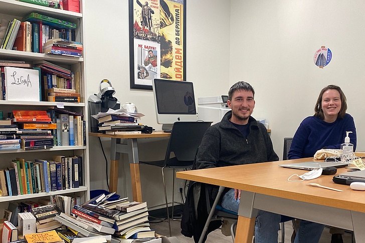 Lächelnder Mann mit grauem Haar in dunkler Jacke und Frau mit Brille in blauem Pullover sitzen an einem vollgestellten Schreibtisch mit Büchern, Monitor, Laptops und Desinfektionsmittel in einem Büro mit Filmplakat an der Wand.
