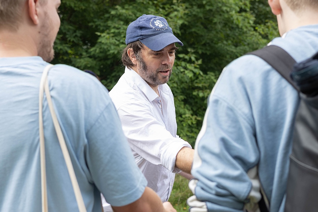 Close-up of a bearded man wearing a cap, handing something to someone. Two men wearing blue T-shirts are seen from behind.