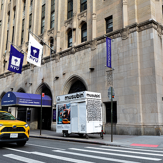 Yellow taxi passes NYU's Frederick Loewe Theatre, with a Musubin kiosk on the sidewalk.
