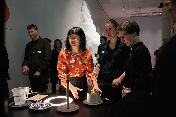 Besucher*innen stehen vor einem dunklen Tisch, auf dem von links nach rechts ein transparentfarbener Plastikeimer, Kabel und ein zylinderförmiges Objekt zu sehen sind.