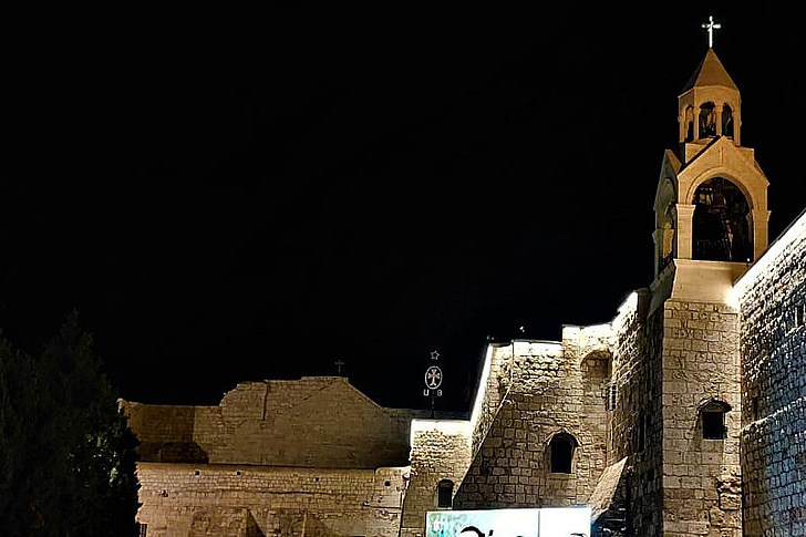 Beleuchtete Steinmauer und Kirchturm mit Glockenturm und Kreuz bei Nacht vor dunklem Himmel