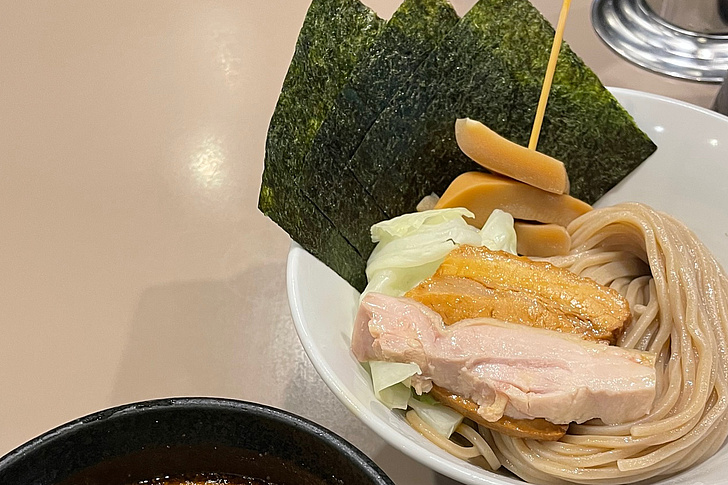Schale mit Soba-Nudeln, belegt mit Hähnchenfleisch, Gemüse, Pilzen und Nori-Blättern, daneben eine Schale mit dunkler Brühe.