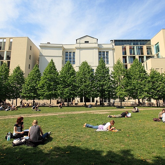 Students relaxing on a green lawn in front of a modern university building on a sunny day.
