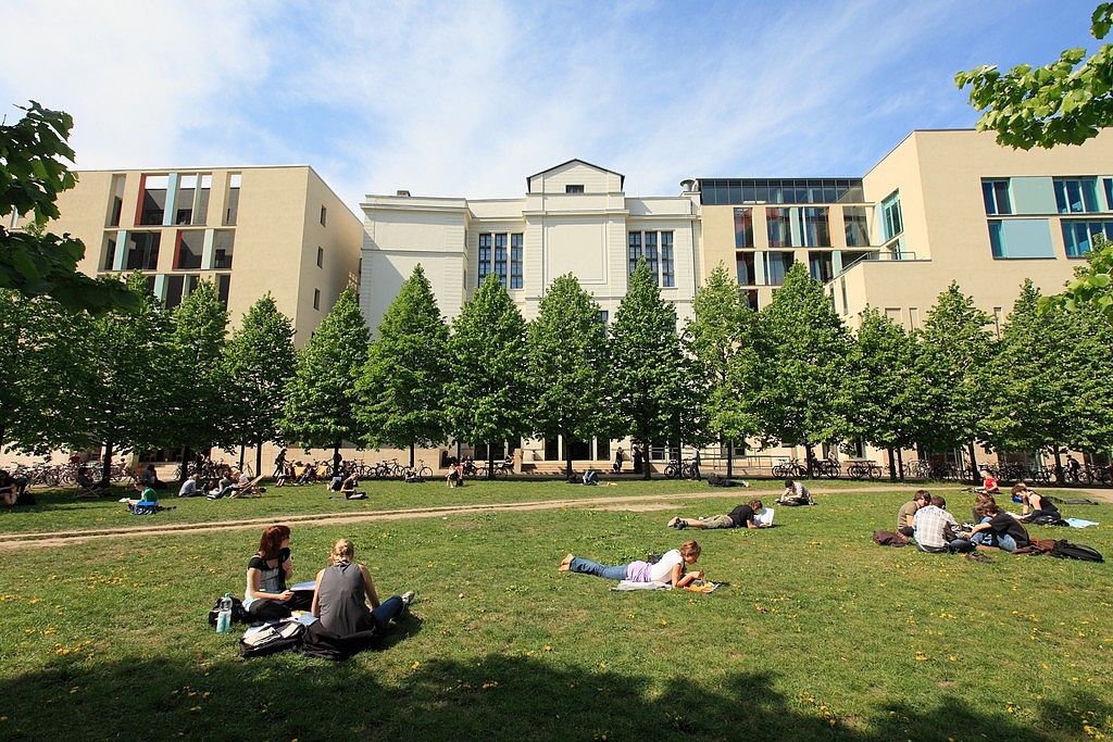 Studierende entspannen auf einer grünen Wiese vor einem modernen Universitätsgebäude bei sonnigem Wetter.