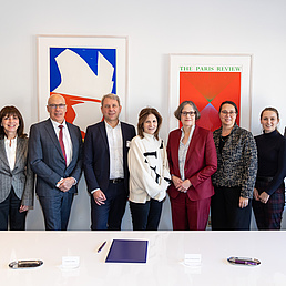26-0307 Group photo of representatives from HU and NYU behind a white table and in front of a white wall with paintings