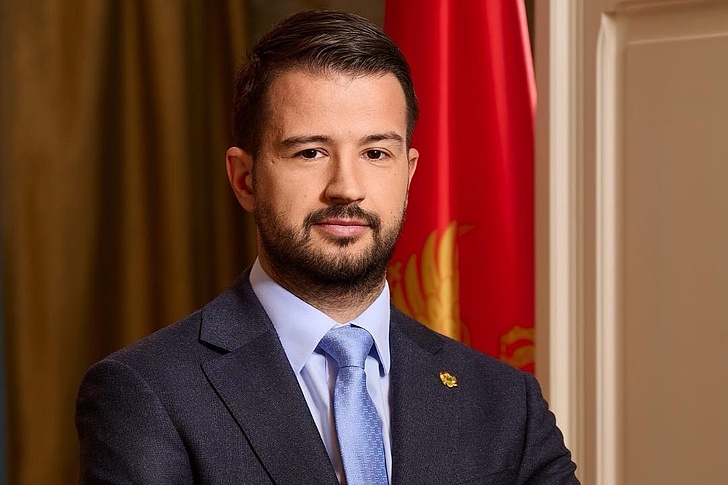 Präsident Milatovic steht aufrecht vor einer Flagge Montenegros. Er hat die Arme überkreuzt und blickt direkt in die Kamera.