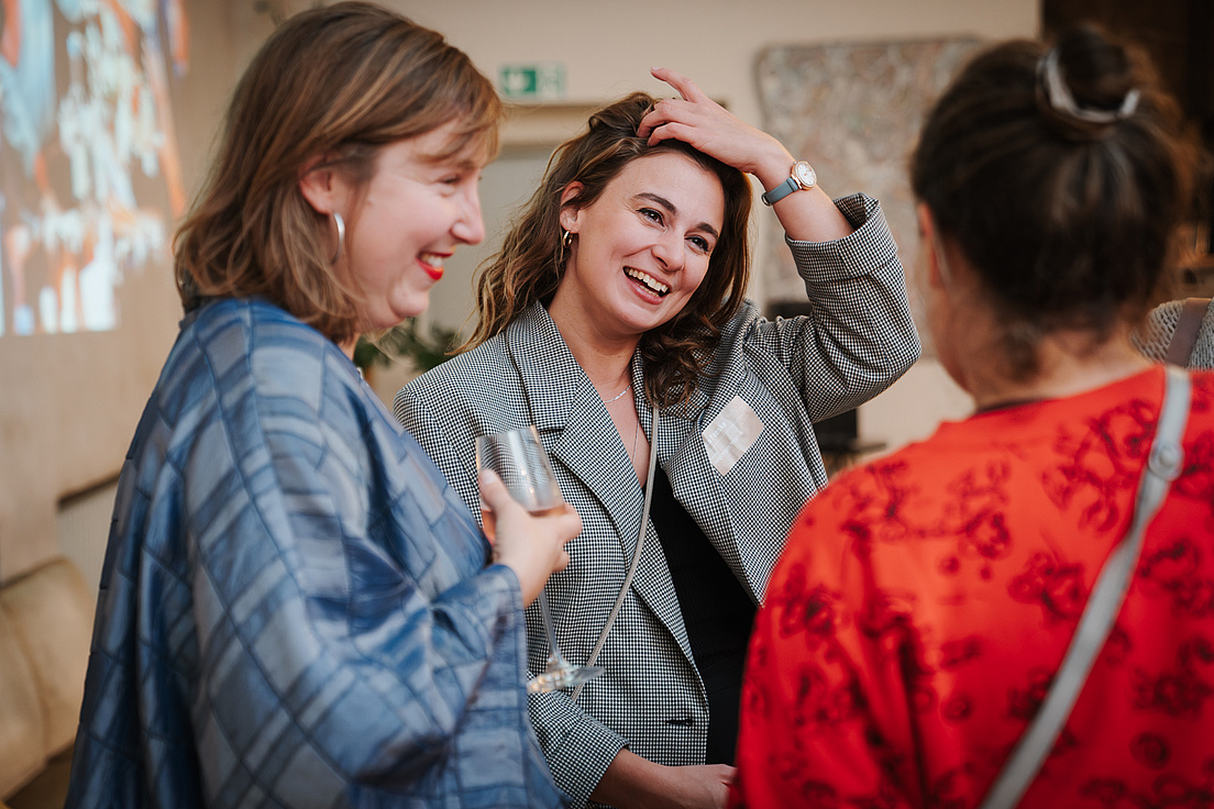 Drei Frauen unterhalten sich bei einer Veranstaltung. Die Frau in der Mitte, in einer karierten Jacke, lacht herzlich und berührt ihr Haar. Die Frau links von ihr, in einer blauen Jacke, hält ein Glas und lacht ebenfalls. Die dritte Frau, rechts im Bild, trägt ein rotes Oberteil mit einem Muster und ist dem Gespräch zugewandt. Im Hintergrund ist eine Projektion zu sehen.