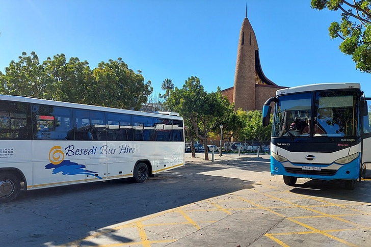 Zwei Shuttlebusse auf einem Parkplatz vor einem Gebäude mit einem hohen, spitzen Turm und Bäumen im Hintergrund