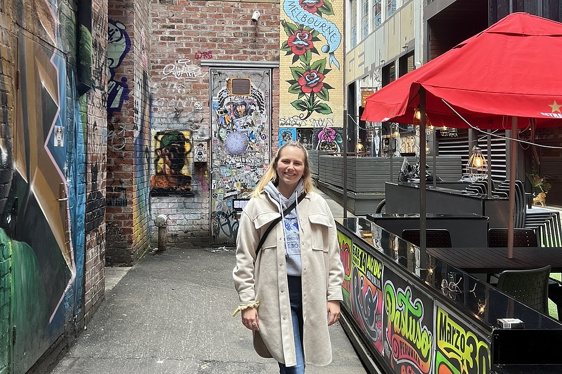 Blonde Frau in beige Daunenjacke und Jeans spaziert grinsend durch eine urbane Gasse mit Streetart-Wänden (Rosenmotive, Graffiti), rotem Schirm und Café-Tischchen.