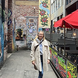 Blonde Frau in beige Daunenjacke und Jeans spaziert grinsend durch eine urbane Gasse mit Streetart-Wänden (Rosenmotive, Graffiti), rotem Schirm und Café-Tischchen.