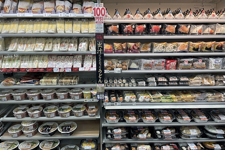 Regal mit verschiedenen verpackten japanischen Lebensmitteln wie Onigiri, Sushi und Fertiggerichten in einem Geschäft.