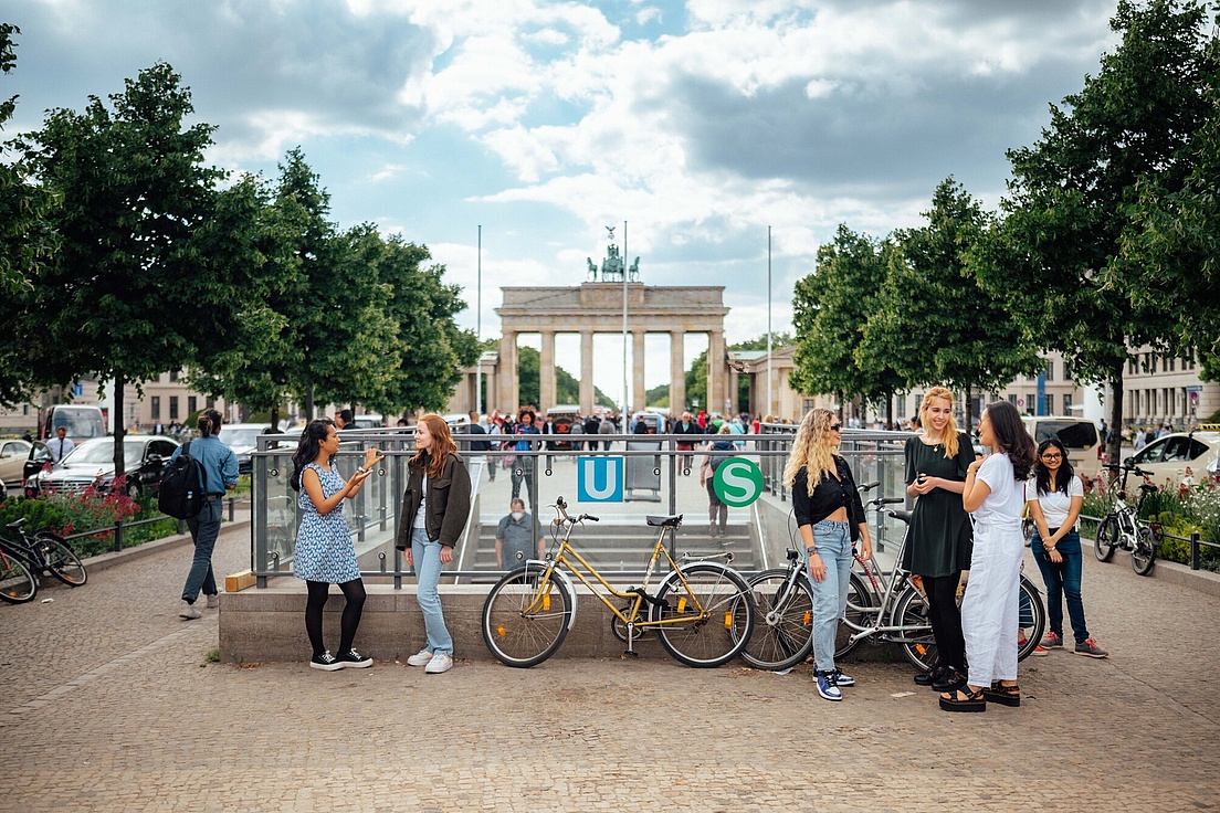 Many people at the surface of a subway station, with cars and other people on the sides, and the Brandenburger Tor visible in the background.