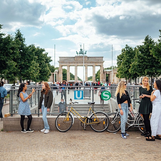 Ganz viele Personen an der Oberfläche einer U-Bahn-Station, an den Seiten stehen Autos und andere Menschen, im Hintergrund sieht man das Brandenburger Tor.
