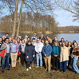 Eine Gruppe von Menschen posiert lächelnd am Ufer eines Sees vor kahlen Bäumen.