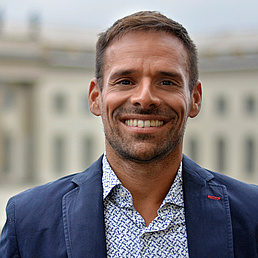 Yoan Vilain ist im Freien und lächelt in die Kamera und trägt ein kornblaues Hemd und ein blau-weiß gemustertes Hemd. Im Hintergrund ist das HU-Hauptgebäude zu sehen.