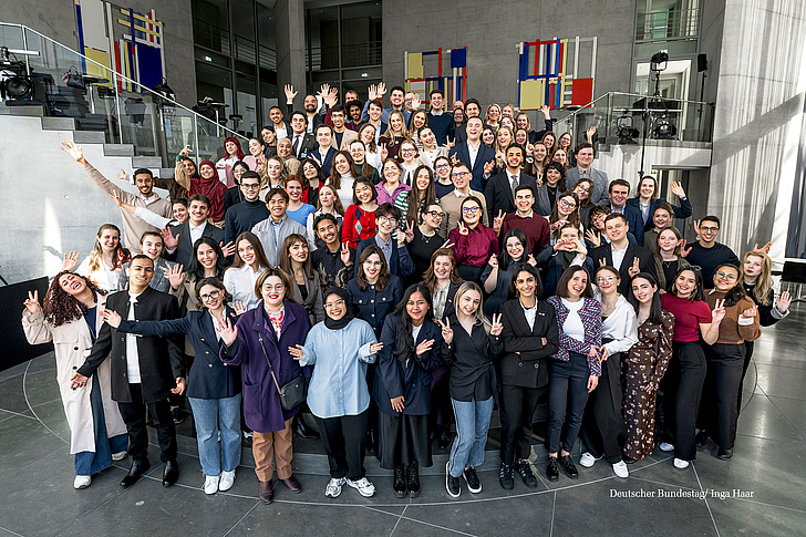 Eine große Gruppe junger Menschen posiert lächelnd in einem modernen Gebäude mit farbigen Kunstwerken an der Wand.