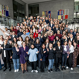 IPS-Stipendiaten Eine große Gruppe junger Menschen posiert lächelnd in einem modernen Gebäude mit farbigen Kunstwerken an der Wand.
