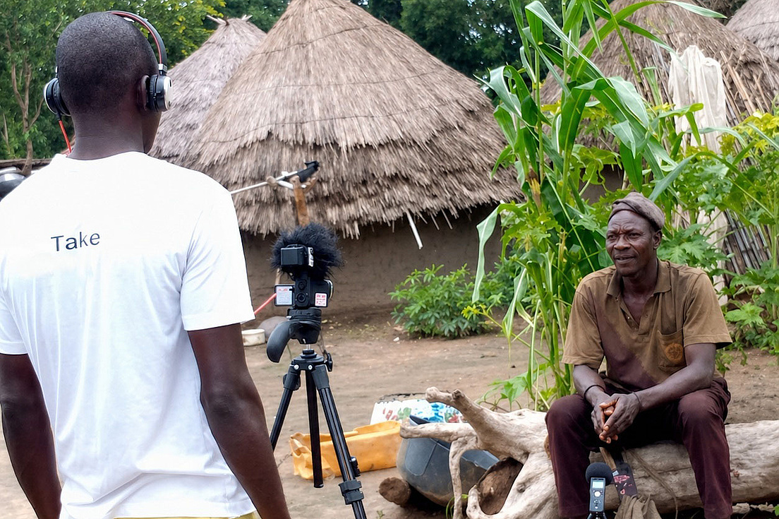 A man is filming an elderly man in a village with thatched roofs.