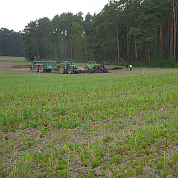 Zu sehen ist ein Feld im Hintergrund ist eine landwirtschaftliche Maschine zu sehen, die den Dünger aus menschlichen Ausscheidungen auf dem Feld verteilt. Im Hintergrund ist der Anfang eines Waldes zu sehen. 