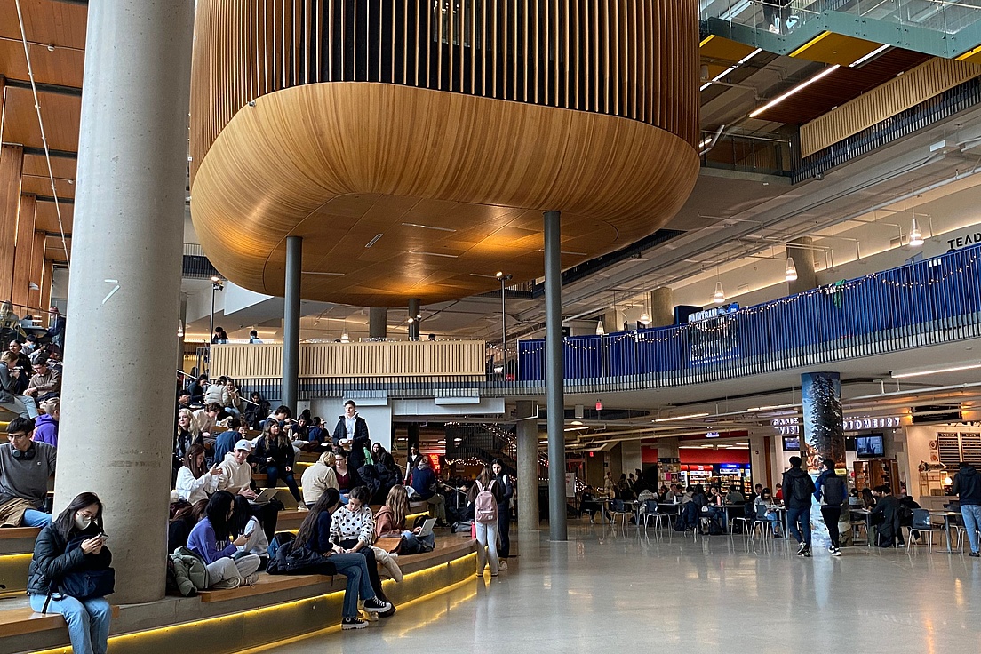 Deutsch: Belebtes modernes Atrium einer Universität oder Einkaufszentrum mit Menschen auf Holzbänken unter einer ovalen Holzpodestkonstruktion an Stahlpfeilern, umgeben von Geschäften und Treppen.