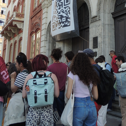 A diverse group of people stands in line outside a building with "ФИЛОЗОФСКИ ФАКУЛТЕТ" inscribed on it, under a large QR code banner.