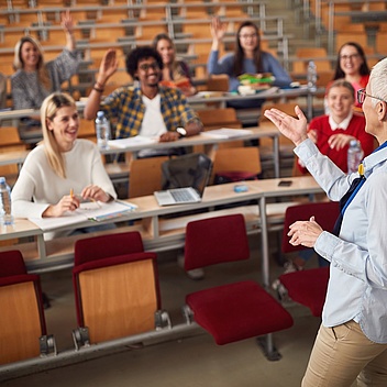 Eine Dozentin spricht vor einer Gruppe von Studierenden in einem Hörsaal. Einige Studierende sitzen in Reihen und heben lächelnd die Hand, während andere Notizen machen oder zuhören.