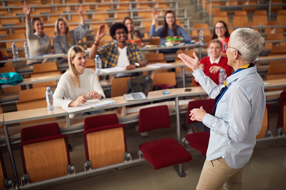Eine Dozentin spricht vor einer Gruppe von Studierenden in einem Hörsaal. Einige Studierende sitzen in Reihen und heben lächelnd die Hand, während andere Notizen machen oder zuhören.