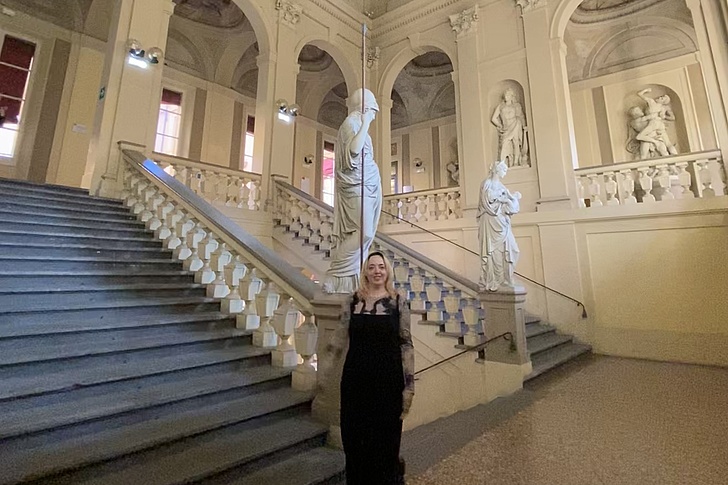 Person in schwarzem Kleid steht vor einer Treppe mit Balustrade und mehreren Statuen in einem großen, hellen Innenraum mit hohen Fenstern und Rundbögen.