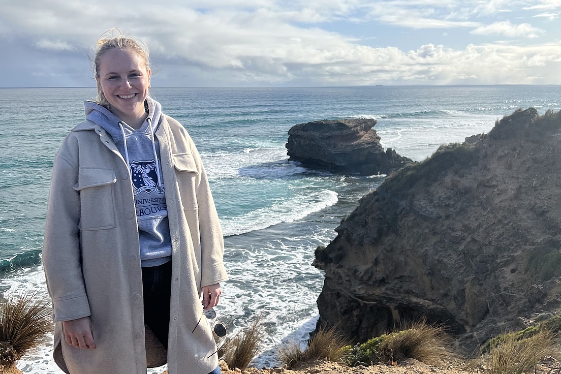 Blonde Frau mit Pferdeschwanz in beige Jacke und blauer World-Hoodie lächelt freudig auf Klippen über türkisfarbenem Ozean mit Felsen und Wellen bei bewölktem Himmel.