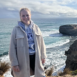 Blonde Frau mit Pferdeschwanz in beige Jacke und blauer World-Hoodie lächelt freudig auf Klippen über türkisfarbenem Ozean mit Felsen und Wellen bei bewölktem Himmel.