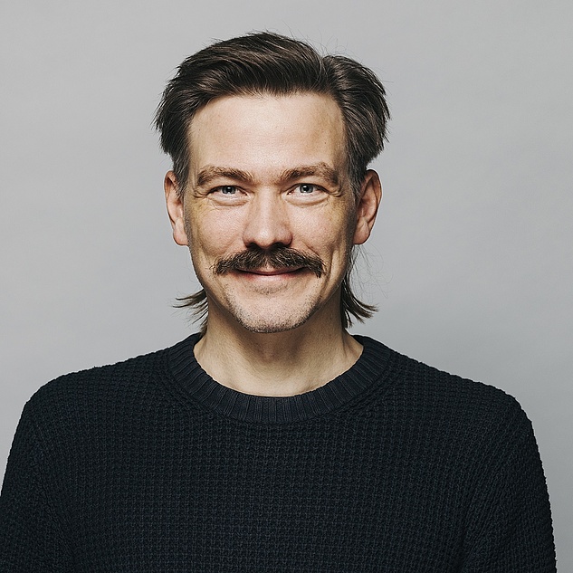 Tim Schröder mit einem Schnurrbart und dunklen Haaren, die nach hinten gekämmt sind, blickt freundlich in die Kamera. Er trägt einen dunklen, gestrickten Pullover und hat ein leichtes Lächeln auf den Lippen. Der Hintergrund ist einheitlich grau.