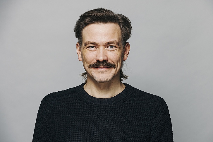 Tim Schröder mit einem Schnurrbart und dunklen Haaren, die nach hinten gekämmt sind, blickt freundlich in die Kamera. Er trägt einen dunklen, gestrickten Pullover und hat ein leichtes Lächeln auf den Lippen. Der Hintergrund ist einheitlich grau.